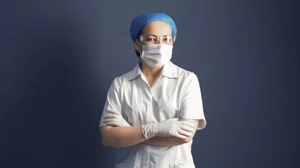 Femeia se uită la brațele încrucișate ale camerei. Femeie medic în mască și uniformă albă, portret pe fundal gri — Fotografie, imagine de stoc