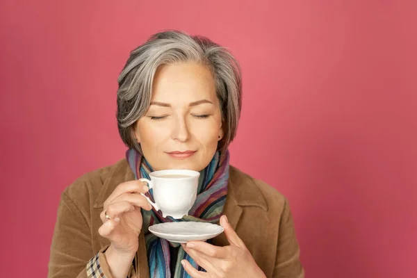 Encantadora mujer adulta bebe café sosteniendo taza blanca y los ojos cerrados. Aislado sobre fondo rosa. Grabado en estudio. concepto de descanso —  Fotos de Stock