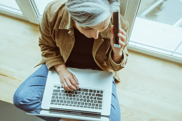 Volwassen vrouw praat mobiele telefoon met laptop computer. Grijs harige blanke vrouw in casual werken zittend gekruiste benen op vensterbank. Hoge hoekaanzicht — Stockfoto