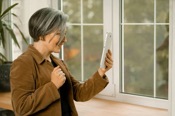 Wanita dewasa cantik berkomunikasi online di tablet digital berdiri di dekat jendela. Smiling woman connecting with friends by internet — Stok Foto