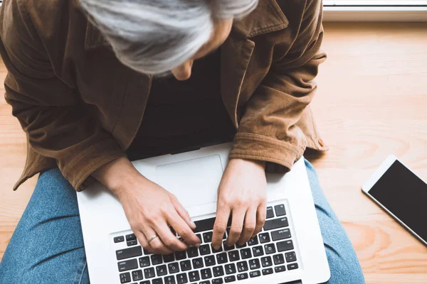 Mulher de cabelos grisalhos digitando teclado laptop sentado no peitoril da janela de madeira com telefone celular nele. Vista superior. Foco seletivo em mãos femininas — Fotografia de Stock