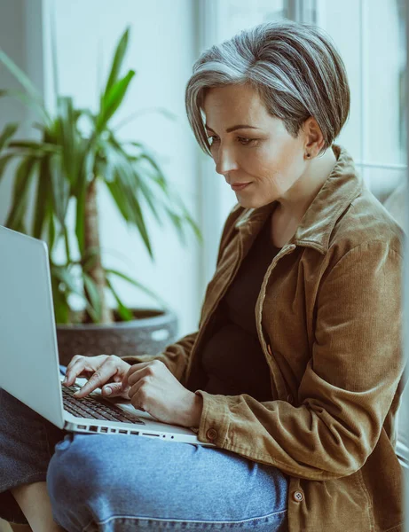 Elegant volwassen vrouw werkt laptop zittend op vensterbank. Zijaanzicht van moderne zilver harige dame in casual typen op computer toetsenbord — Stockfoto