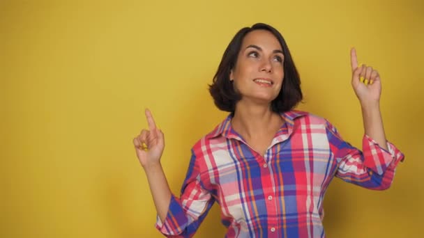 Brunette laat haar wijsvingers zien. Ze probeert een keuze te maken uit twee opties. jonge vrouw glimlacht in studio op gele achtergrond. — Stockvideo