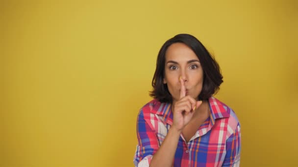 Brunett kvinna ber att vara tystare. Hon sätter fingret på läpparna och ler. Fotografering i studion på en isolerad gul bakgrund — Stockvideo