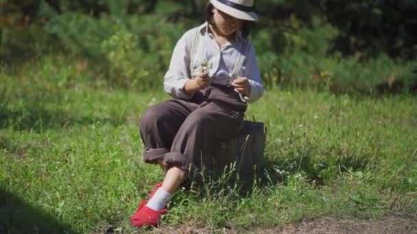 Farmerka zbiera kwiaty łąki siedząc na pniu. Młoda kobieta w kapeluszu i spodniach z szelkami niesie zebrany bukiet innej kobiecie. Wysokiej jakości materiał 4k. 29 sierpnia 2020 r. — Wideo stockowe