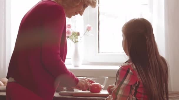 Blond oude vrouw leert bruinharig meisje hoe tomaat l snijden. Kleindochter en oma relatie. Gezinsbevruchting — Stockvideo