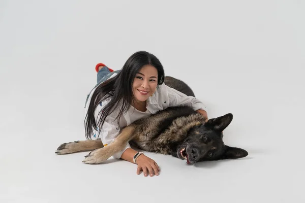 Aziatische vrouw ligt met een herdershond op de studiovloer. Eigenaar knuffelt de hond op een geïsoleerde achtergrond. Vrouw kijkt in de camera en glimlacht. Hoge kwaliteit foto — Stockfoto