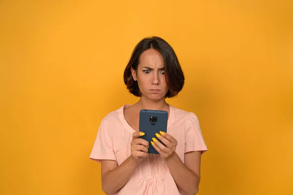 Wanita muda tampak bingung pada ponsel pada latar belakang studio kuning terisolasi. Brunette memegang ponsel. Foto berkualitas tinggi — Stok Foto