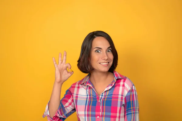 Девушка показывает пальцам знак "хорошо". Брюнетка улыбается и кладет пальцы на кольцо на жёлтом фоне. Высокое качество фото — стоковое фото