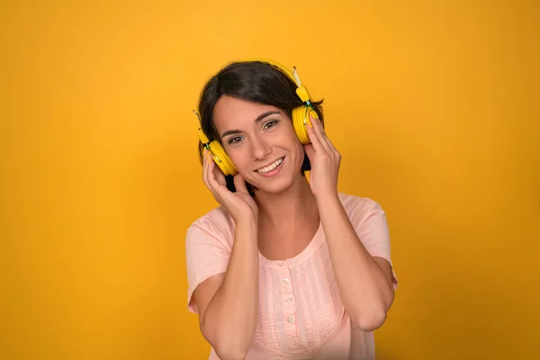 Νεαρή γυναίκα πιέζει κίτρινα ακουστικά στα αυτιά της στο στούντιο. Η Μπρουνέτ χαμογελάει και ακούει μουσική σε κίτρινο φόντο. Υψηλής ποιότητας φωτογραφία — Φωτογραφία Αρχείου