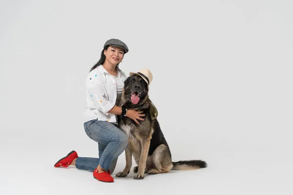 Asian woman in a cap with an Eastern European Shepherd in a hat sits on the floor of the studio. Owner and dog in hats on an isolated background. High quality photo — Stock Photo, Image