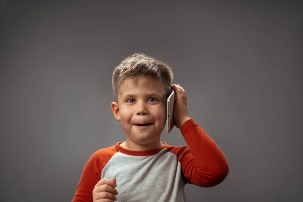 Muchacho divertido sostiene el teléfono móvil o tableta por su oído en el estudio. Retrato de un chico con un artilugio. Aislado sobre fondo gris. Foto de alta calidad — Foto de Stock