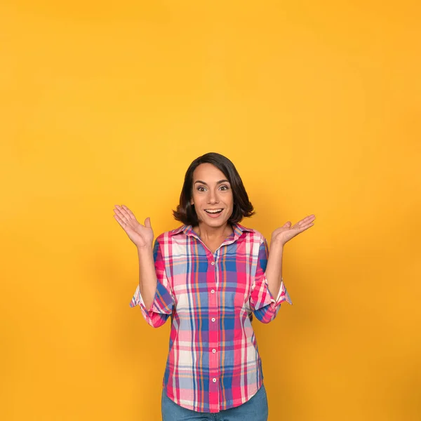 Jonge vrouw heft haar gebogen armen op en haalt haar schouders op. Brunette in geruit shirt in studio op gele achtergrond. Hoge kwaliteit foto — Stockfoto