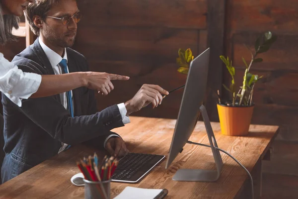 Trabajo en equipo de coworking. El hombre con gafas se sienta en una mesa y apunta a un monitor. Muestra la mano de las mujeres en el monitor de la computadora. Foto de alta calidad — Foto de Stock