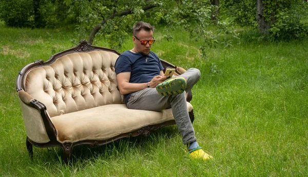 Hombre de mediana edad utiliza el teléfono móvil descansando en el sofá de luz en el jardín. Sofá está en la hierba verde al aire libre. Foto de alta calidad — Foto de Stock