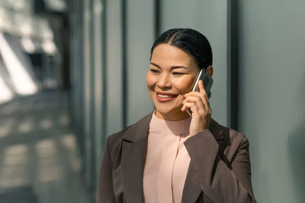 Asiatka mluví na ulici po mobilu. Podnikatelka v bundě drží telefon u ucha a usmívá se. Kvalitní fotografie — Stock fotografie