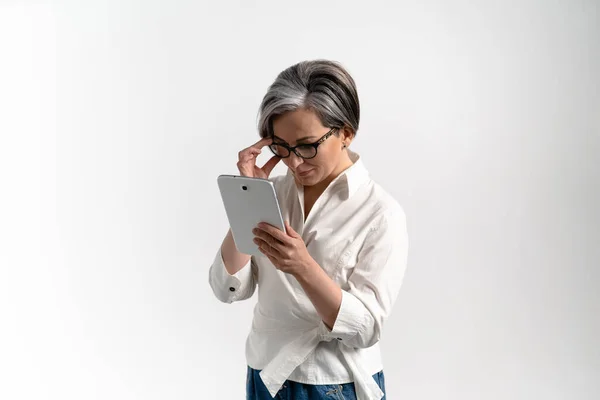 Woman with gray hair in glasses holds a tablet and looks into it. Adult woman on an isolated studio background. High quality photo — Stock Photo, Image
