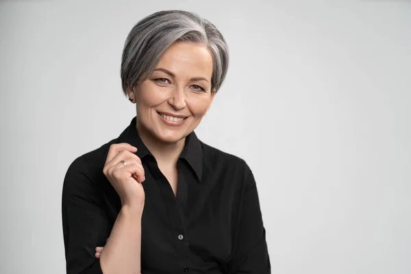 Affascinante donna dai capelli grigi che indossa camicia nera sorride alla macchina fotografica, isolata su sfondo bianco. Maturo donna caucasica in studio alzò il gomito piegando il braccio. Foto di alta qualità — Foto Stock