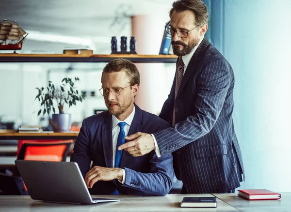 Hombres detrás del portátil en la oficina. Un hombre con traje se sienta en una mesa con un portátil. Su colega está a su lado y señala la pantalla. Foto de alta calidad — Foto de Stock