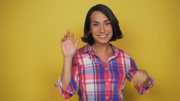 Mujer joven agita los brazos y envía un beso aéreo a la cámara sobre un fondo amarillo. Morena sonríe saludando saludando con la mano. Alta calidad — Vídeos de Stock