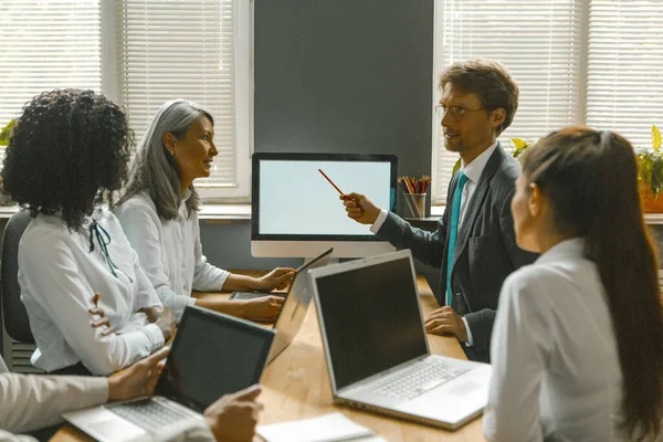 Παρουσίαση του επιχειρηματικού σχεδίου κατά τη διάρκεια συνάντησης διαφόρων ομάδων στο γραφείο. Νεαρός Καυκάσιος άνδρας μοιράζεται ιδέες με γυναίκες συναδέλφους δείχνοντας με μολύβι στη λευκή οθόνη της οθόνης του υπολογιστή — Φωτογραφία Αρχείου