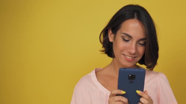 Mulher de cabelos castanhos jovem rolando seu telefone. Modelo sorri enquanto assiste seu telefone no backgr amarelo — Vídeo de Stock