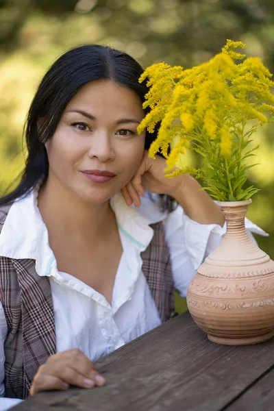 Bellezza donna asiatica con fiori gialli a riposo all'aperto. Giovane bruna si siede a tavolo di legno con bouquet di Solidago o Canada Goldenrod su di esso. Ritratto femminile romantico — Foto Stock