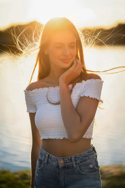 Felice adolescente sorridente con la luce solare retroilluminata al tramonto nella natura. Affascinante ragazza caucasica sullo sfondo del fiume. Concetto vacanza estiva — Foto Stock