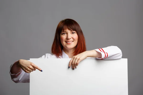 Foto de una chica chef sonriente con un abrigo blanco señalando con el dedo la hoja vacía para la inscripción — Foto de Stock