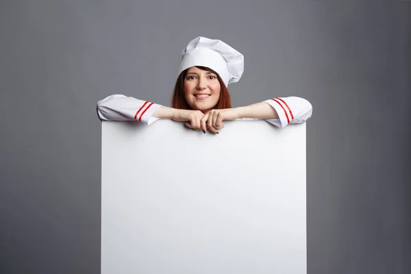 Foto del chef morena sonriente en bata blanca gorra con papel vacío en las manos — Foto de Stock