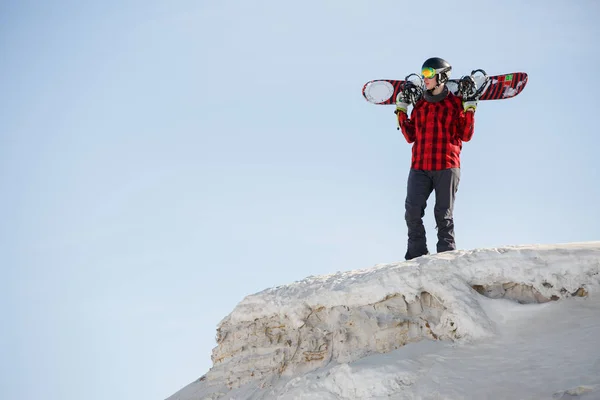 Foto de hombre en casco con tabla de snowboard sobre hombros de pie en la ladera de la montaña nevada —  Fotos de Stock