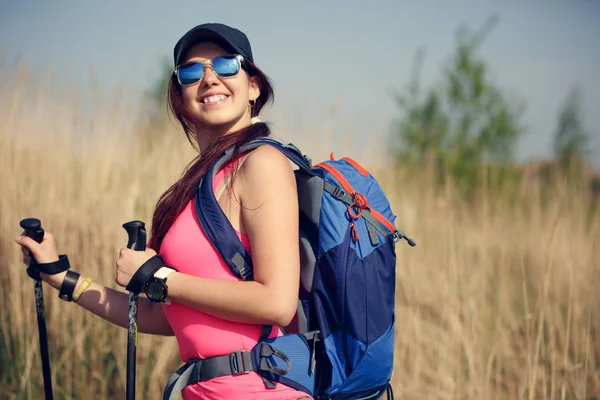 Foto de hermosa chica turística con mochila y bastones en el fondo del campo y el cielo —  Fotos de Stock