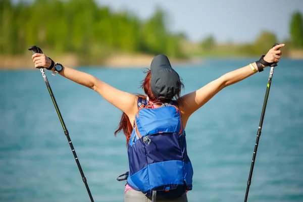 Photo of back of sporty woman with walking sticks and backpack on lake background — Stock Photo, Image