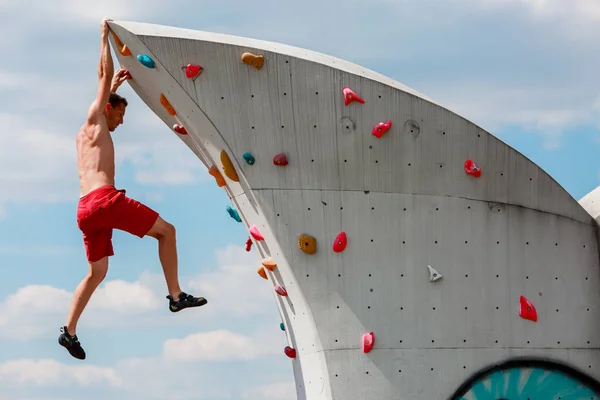 Zdjęcie młodych sportowy człowieka w czerwonych spodenkach, wiszące na ścianie dla wspinaczki przeciw błękitne niebo z chmurami — Zdjęcie stockowe