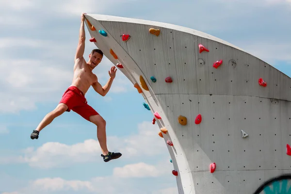 Foto de jovem desportista em shorts vermelhos pendurados na parede para escalada de rocha contra o céu azul com nuvens — Fotografia de Stock