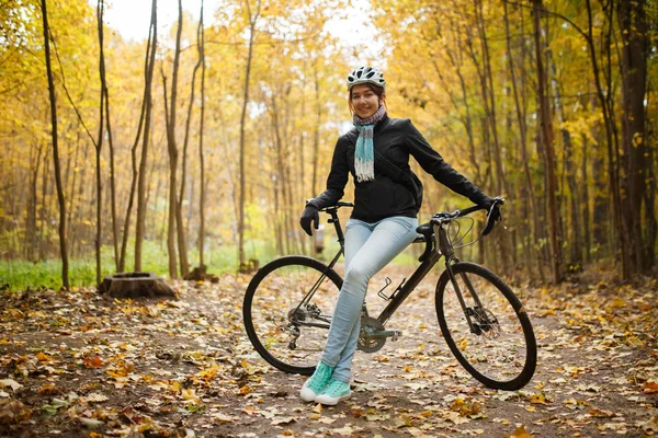 Foto de morena en casco, jeans al lado de la bicicleta en el parque de otoño —  Fotos de Stock