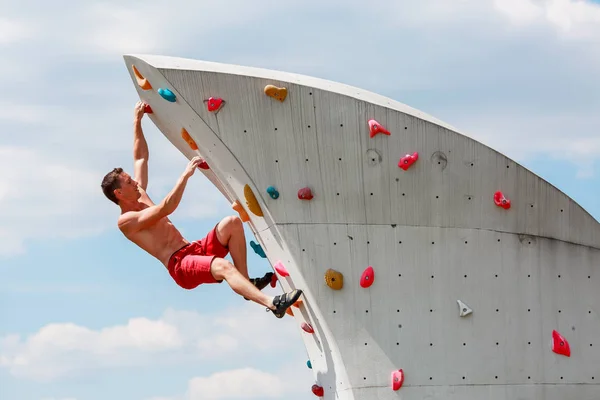 Photo of young sporty man in red shorts hanging on wall for rock climbing against blue sky with clouds — Stock Photo, Image