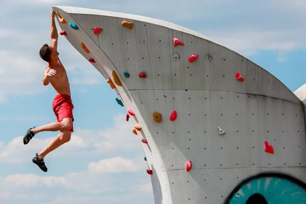 Foto eines starken Sportlers in roten Shorts, der an der Wand hängt, um gegen den blauen Himmel mit Wolken zu klettern — Stockfoto