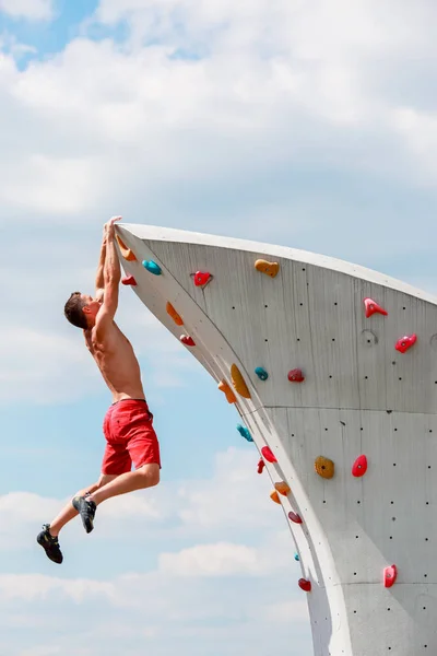 Foto eines jungen sportlichen Mannes in roten Shorts, der an der Wand hängt, um gegen den blauen Himmel mit Wolken zu klettern — Stockfoto