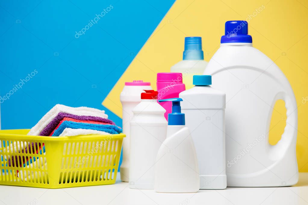 Photo of bottles of cleaning products and multi-colored towels in basket isolated on yellow,blue background