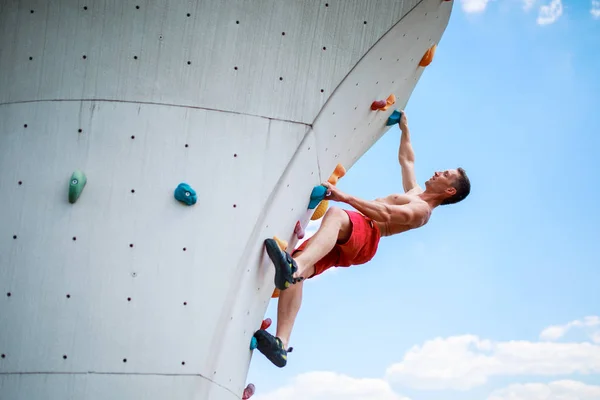 Foto von unten von starkem athletischen Kerl, der an der Wand übt, um gegen blauen Himmel mit Wolken zu klettern — Stockfoto