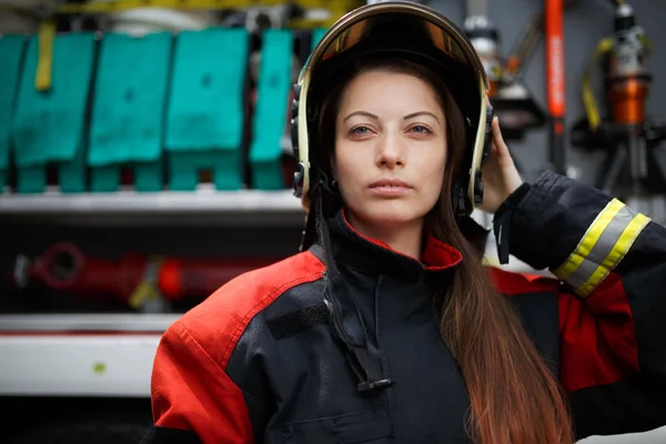 Foto de una joven bombera con el pelo largo en el casco mirando a la cámara junto al camión de bomberos — Foto de Stock
