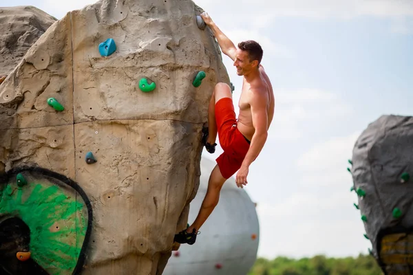 Foto eines lächelnden Typen in roten Shorts, der auf Felsbrocken übt, um gegen bewölkten Himmel zu klettern — Stockfoto