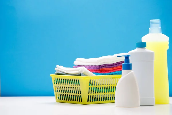 Fotografie ze tří lahví pro čisticí prostředky a žlutý koš s ručníky na prázdné modré pozadí — Stock fotografie