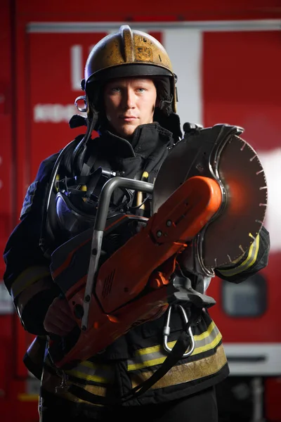 Photo of serious fireman wearing helmet with chainsaw on background of fire engine