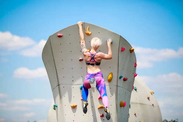 Foto von hinten von jungen athletischen Blondine in Leggings hängen an der Wand zum Klettern gegen den blauen Himmel — Stockfoto
