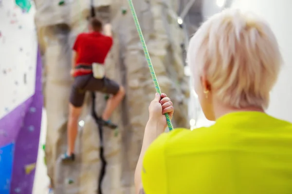 Photo from back of man clambering over climbing wall and female trainer holding safety rope in gym — Stock Photo, Image