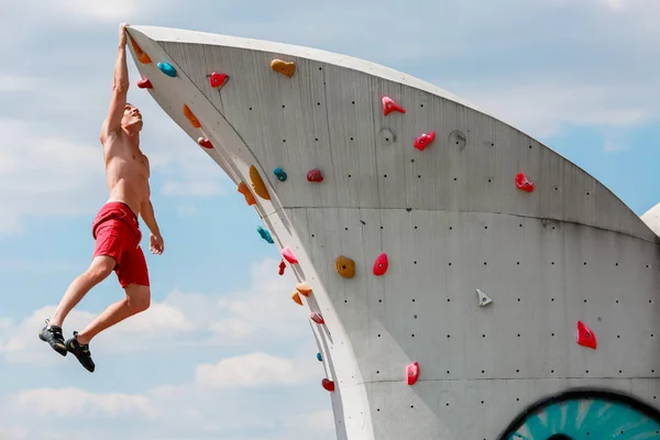 Photo of trained sports man in red shorts hanging on wall for rock climbing against blue sky with clouds