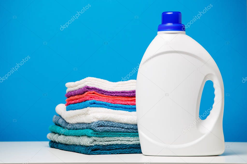 Photo of one white bottle of cleaning agent and stack of multi-colored towels isolated on blue background