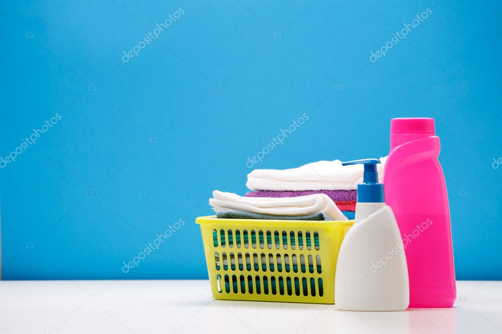 Photo of pink and white bottle for cleaning products and yellow basket with towels on empty blue background